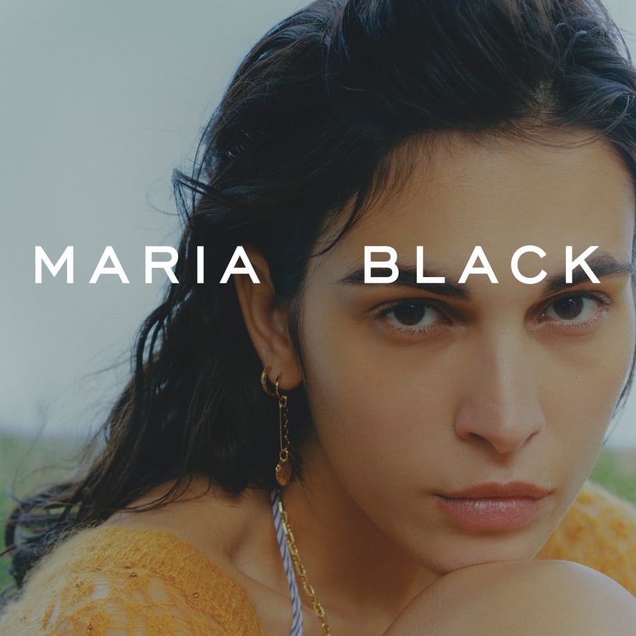 Mørkhåret ung kvinde der kigger ind i kameraet og har en halskæde og øreringe på af mærket Maria Black. På billedet er der en tekst i hvid hvor der står Maria Black.